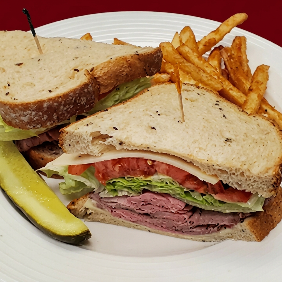 Two deli sandwiches with lettuce, tomato, and sliced meat served with French fries and a pickle on a white plate.